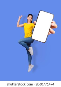 Full length of smiling young Asian woman jumping, gesturing peace and showing mobile phone with empty screen on blue studio background, collage with mockup for app or website - Shutterstock ID 2060771474