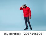 Full length of smiling young bearded african american man 20s wearing casual red shirt hold cap eyeglasses standing and looking camera isolated on pastel blue color wall background studio portrait