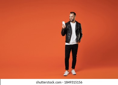 Full length of smiling young bearded man 20s wearing basic white t-shirt black leather jacket standing using mobile cell phone typing sms message isolated on orange colour background studio portrait