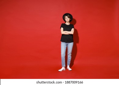 Full length of smiling young brunette woman 20s years old wearing casual basic black t-shirt hat standing holding hands crossed looking camera isolated on bright red color background studio portrait