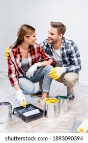 Full Length Of Smiling Young Couple Looking At Each Other While Squatting Near Tins Of Paint, Rollers And Tray At Home