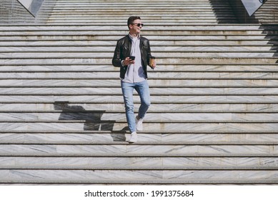 Full Length Smiling Young Man In Black Leather Jacket Glasses Walk In City Downtown Go Down Concrete Steps Hold Mobile Cell Phone Takeaway Craft Paper Cup Drink Coffee To Go Urban Lifestyle Concept