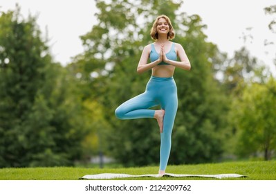 Full length of smiling young sporty girl in sportswear standing outside in Tree Vrksasana pose against background of green lush trees in city park, female during yoga class in nature - Powered by Shutterstock