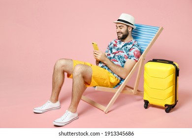 Full length smiling young traveler tourist man in hat sit on deck chair using mobile cell phone typing sms message isolated on pink background. Passenger travel on weekend. Air flight journey concept