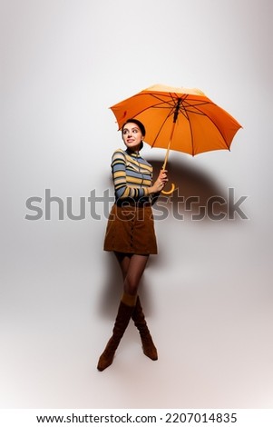full length of smiling young woman in striped turtleneck and skirt posing with orange umbrella on grey