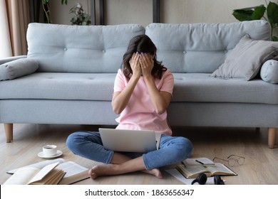 Full length stressed millennial mixed race female student sitting on floor with books and computer on laps, feeling under pressure exhausted preparing for university examinations alone at home.