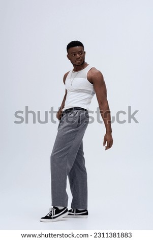 Full length of strong black man in pants and tank top standing and posing confidently in stylish and trendy outfit on grey background
