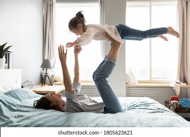 Full Length Strong Young Smiling Mommy Lying On Comfortable Bed, Holding Small Kid Daughter On Straight Legs. Happy Little Child Girl Flying In Air, Practicing Yoga Activity With Mother In Bedroom.