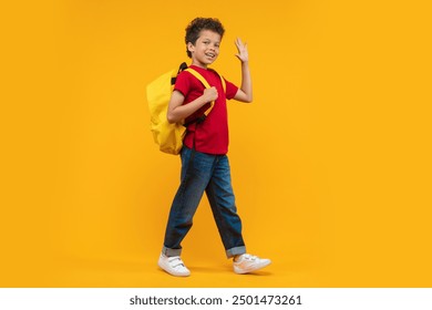 Full length studio portrait of cute casually dressed confident African American schoolboy walking with his bright colored backpack looking to the camera and waving hand, isolated on yellow background - Powered by Shutterstock