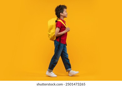 Full length studio portrait of a cute casually dressed happy smiling confident African American schoolboy walking with his bright colored backpack over orange yellow background. - Powered by Shutterstock