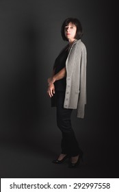 Full Length Studio Portrait Of An Older Woman Standing With Hands Folded, Sweater Over Her Shoulder, As If She Is Looking The Viewer Over.