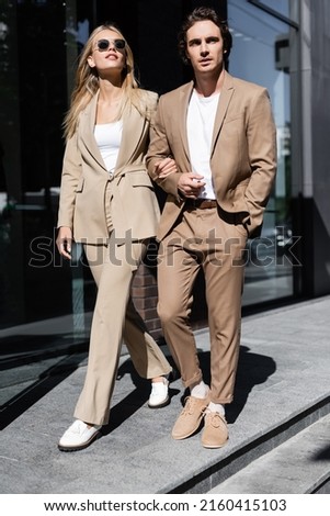 full length of stylish couple in beige suits walking on urban street