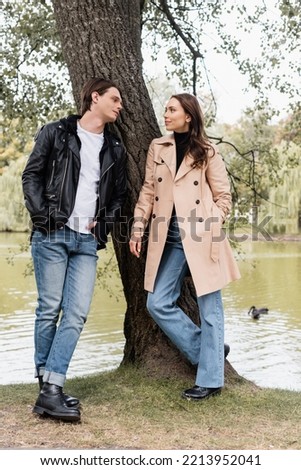 full length of stylish man and pretty woman in trench coat looking at each other near river in park