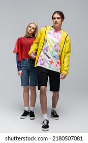 Full Length Of Stylish Man In Windbreaker Holding Hand Of Girlfriend On Grey Background