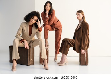 stylische multikulturelle Frauen in Anzügen, die auf Koffer auf Weiß sitzen