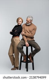 Full Length Of Stylish Woman Crossing Arms Near Mature Husband Sitting On Chair On Grey Background