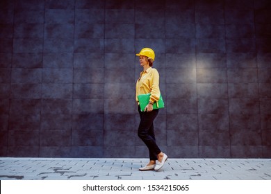 Full Length Of Successful Caucasian Blonde Senior Female Architect With Helmet On Head And Folder With Documents Under Armpit Passing By Gray Wall And Going To The Construction Site.