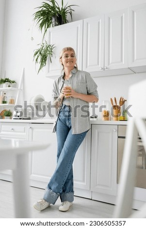 full length of tattooed and happy woman in eyeglasses holding glass of orange juice near kitchen worktop with clean dishes, toaster and rack with plants looking at camera in modern apartment