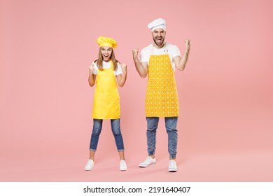 Full Length Teen Fun Girl Dad Man Father's Helper Chef Cook Confectioner Baker In Yellow Apron Toque Cap Doing Winner Gesture Celebrating Clench Fist Isolated On Pastel Pink Background Studio Portrait