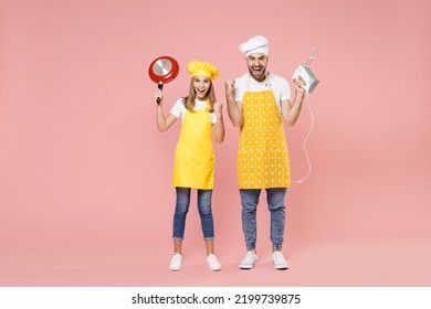 Full Length Teen Fun Girl Dad Father's Helper Chef Cook Confectioner Baker In Yellow Apron Cap Hold Mixer Blender Frying Pan Do Winner Gesture Clench Fist Isolated On Pink Background Studio Portrait