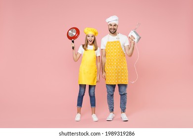 Full Length Teen Satisfied Happy Fun Girl Dad Father's Helper Chef Cook Confectioner Baker Wearing Yellow Apron Cap Holding Mixer Blender Frying Pan Isolated On Pastel Pink Background Studio Portrait