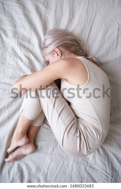 Full length top above view stressed middle aged\
woman lying on bed in fetal position, embracing knees, suffering\
from depression or insomnia. Upset mature lady feeling unwell,\
having health problems.