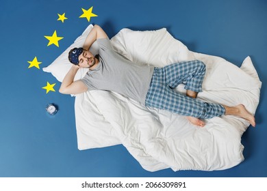 Full length top view calm young caucasian man in pajamas jam sleep mask rest relax at home lies in bed hold hands behind head neck isolated on dark blue sky background Good mood night bedtime concept