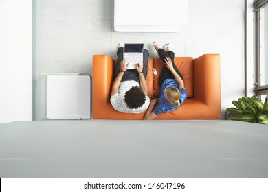 Full Length Top View Of A Man And Woman Using Laptop On Orange Sofa In Reception Room At Office