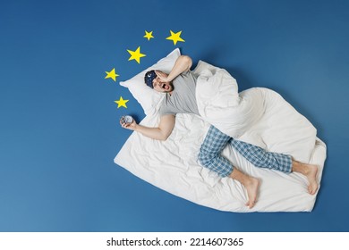 Full length top view young latecomer man in pajamas jam sleep mask rest at home lie wrap covered under blanket hold face slept late isolated on dark blue sky background Bad mood night bedtime concept