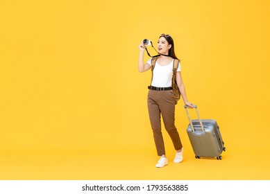 Full Length Travel Portrait Of Excited Young Tourist Asian Woman Walking While Taking Photo With Camera In Isolated Studio Yellow Background