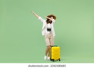 Full length traveler tourist woman in casual clothes hat hold suitcase doing dab hip hop dance gesture isolated on pastel green background. Passenger travel abroad weekends. Air flight journey concept