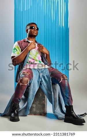 Full length of trendy african american model in sunglasses, colorful denim vest and t-shirt sitting on stone on grey with blue polycarbonate sheet at background, fashion shoot, DIY clothing