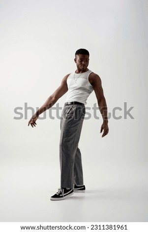 Full length of trendy young african american man in pants, sleeveless t-shirt and sneakers standing on grey background, contemporary shoot featuring casual attire