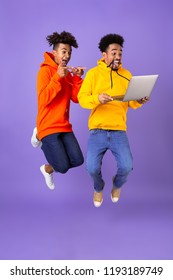 Full Length Of Two Happy Male African Friends In Colorful Hoodies Jumping Isolated Over Violet Background, Holding Laptop Computer