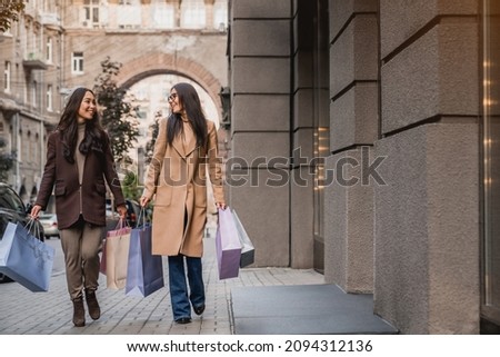 Full length of two young women girlfriends walking on the city street with shopping bags after buying purchasing fashion clothes, Christmas presents on sales offers discounts
