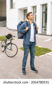 Full Length Vertical Portrait Of Cheerful Handsome Young Delivery Man With Thermo Backpack Standing On City Street, On Background Of Bicycle And Office Building, Looking Up.