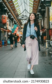 Full Length With Vertical Shot Of Relaxed Asian Female Traveler With Camera Looking Into Distance While Exploring Inside Local Kuromon Ichiba Market In Osaka Japan