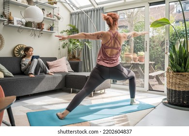 Full Length View Of The Adult Woman Wearing Home Wear Practicing Yoga In Warrior Pose At The Light Living Room While Her Daughter Sitting At The Sofa 