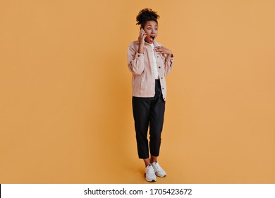 Full Length View Of Black Woman Talking On Smartphone. Studio Shot Of African American Girl In Sneakers Standing On Orange Background.