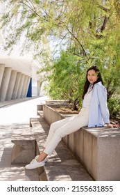 Full Length View Of Brunette Woman In White Pants Sitting Under Trees On City Street