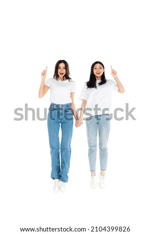 full length view of cheerful multiethnic women pointing with fingers while levitating isolated on white