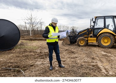 Full length view of construction engineer reviewing project documentation near excavator at outdoor building site. Industrial development and supervision concept.