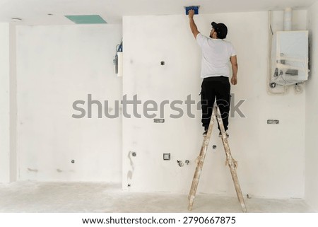Full length view of contractor on ladder performing ceiling finishing work in unfinished home interior, renovation and construction industry concept