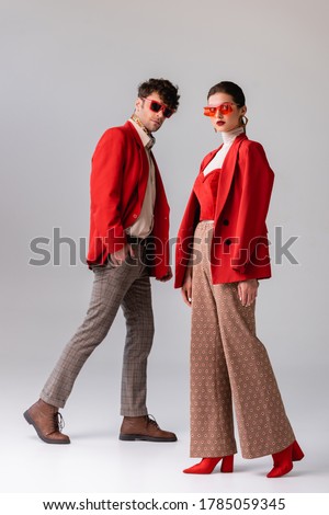 full length view of fashionable couple in red blazers and sunglasses looking at camera  on grey
