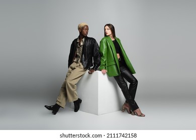 Full Length View Of Fashionable Interracial Couple Sitting On White Cube And Looking At Each Other On Grey