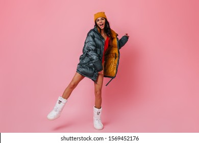 Full length view of girl in down jacket and winter boots. Studio shot of blissful brunette woman dancing on pink background.