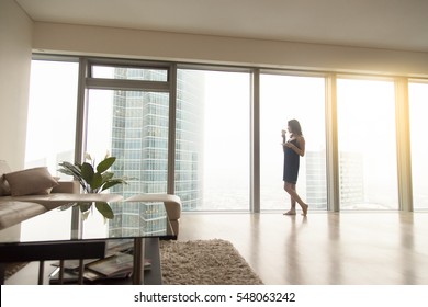 Full Length View Of Happy Young Woman Model Standing Near The Window With A Cup Of Coffee In Luxury Modern Apartment, Looking At Cityscape After Waking Up In The Morning, Relaxing, Dreaming