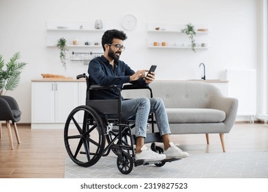 Full length view of indian male with disability using smartphone while resting in modern kitchen of apartment. Joyful person in glasses and casual clothes texting message on digital device at home. - Powered by Shutterstock