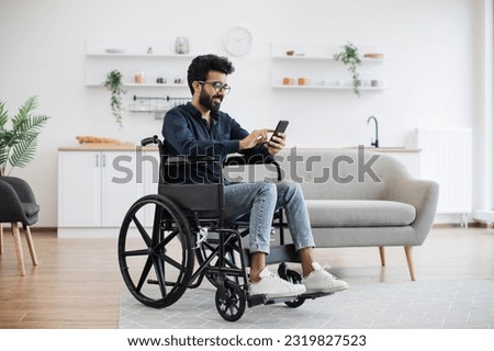 Full length view of indian male with disability using smartphone while resting in modern kitchen of apartment. Joyful person in glasses and casual clothes texting message on digital device at home.