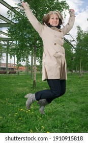 Full length view of a mature woman in a trench coat in a public park with arms up, jumping joyfully. 